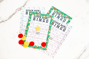 Four printed Super Bowl Bingo cards&mdash;two color and two black-and-white&mdash;layered on a marble surface with red and yellow bingo markers on top.
