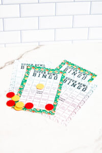Three Super Bowl Bingo sheets, both color and black-and-white versions, arranged on a marble surface with bingo markers placed on several squares.