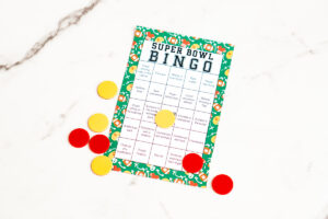 Single colorful Super Bowl Bingo card on a marble surface with several yellow and red bingo markers positioned around and on the card.