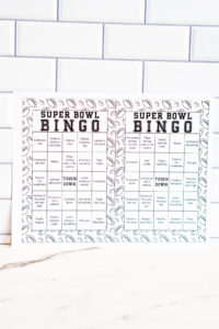 Close-up of two black-and-white Super Bowl Bingo cards with football-themed doodle borders displayed against a subway tile background.