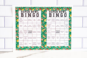 Two colorful Super Bowl Bingo cards with green illustrated borders laid side by side on a white tiled background.