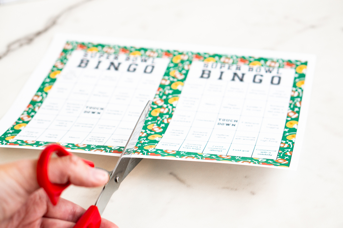 Hand holding red scissors, trimming the edge of a printed green-bordered Super Bowl Bingo sheet on a white surface.