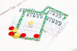 Assorted printed Super Bowl Bingo cards spread out on a white surface with red and yellow bingo markers placed on top.