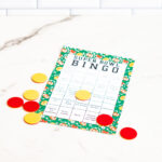 Single printed Super Bowl Bingo card with a green football-themed border on a marble surface, surrounded by scattered red and yellow bingo markers.