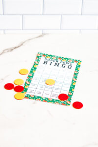 Single printed Super Bowl Bingo card with a green football-themed border on a marble surface, surrounded by scattered red and yellow bingo markers.