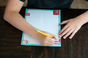 Child writing on a colorful Dear Santa letter printable with a pencil at a wooden table.
