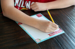 Child writing on a colorful Dear Santa letter printable with a pencil at a wooden table.