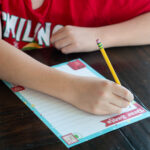 Child writing on a colorful Dear Santa letter printable with a pencil at a wooden table.