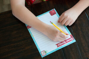 Child writing on a colorful Dear Santa letter printable with a pencil at a wooden table.