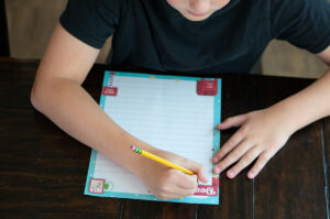 Child writing on a colorful Dear Santa letter printable with a pencil at a wooden table.