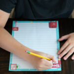 Child writing on a colorful Dear Santa letter printable with a pencil at a wooden table.