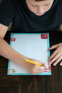 Child writing on a colorful Dear Santa letter printable with a pencil at a wooden table.