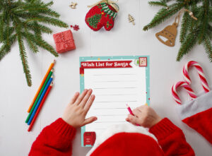 Child wearing a red sweater fills out a Wish List for Santa printable surrounded by Christmas decorations, candy canes, colored pencils, and evergreen branches.