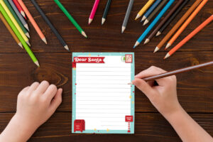 Child&rsquo;s hands writing on a Dear Santa letter printable surrounded by scattered colored pencils on a dark wood table.