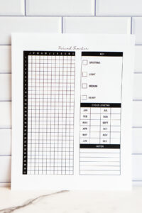 Printed yearly period tracker page displayed against a white subway tile backdrop. The page includes a vertical 31-day grid labeled with months, a symptom key for spotting through heavy flow, a cycle length chart for each month, and a lined notes section.