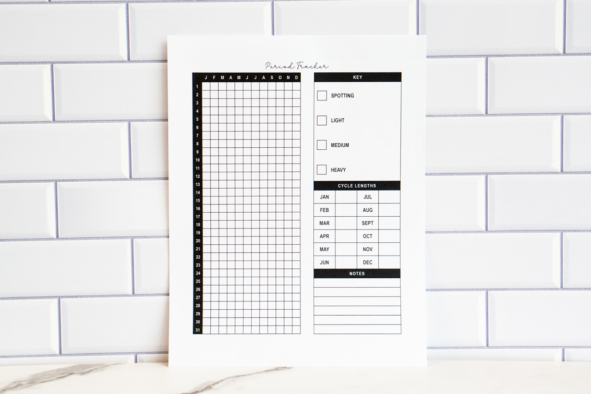 Printed yearly period tracker page displayed against a white subway tile backdrop. The page includes a vertical 31-day grid labeled with months, a symptom key for spotting through heavy flow, a cycle length chart for each month, and a lined notes section.