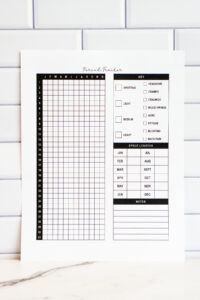 Yearly period tracker printable displayed upright against a white subway tile background. The page features a 31-day vertical grid labeled with months, a detailed symptom key including spotting, cramps, cravings, mood swings, fatigue, and more, a cycle length chart for each month, and a lined notes section at the bottom.