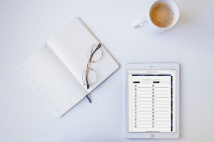Flat lay photo of an open lined planner with glasses resting on the pages, a cup of coffee, and an iPad displaying an alphabet reading challenge printable on a white desk.