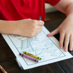 Child wearing a red shirt coloring a printable relaxation coloring page at a table, with crayons scattered nearby.