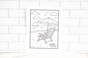 Full view of the beach-themed coloring page showing a beach chair under an umbrella at sunset, displayed against a white tile wall.