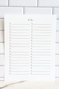 Photo of a simple two-column printable to-do list with checkboxes and lined sections, shown in portrait orientation against a white subway tile background.