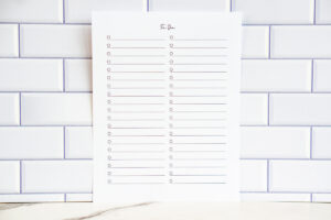 Full view of a clean black-and-white two-column printable to-do list page with checkboxes, displayed upright in front of white subway tile.