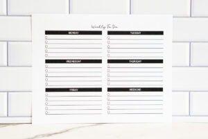 Printable weekly to-do list page in landscape orientation with sections for Monday through Weekend, photographed against white subway tile.