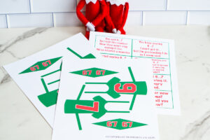 Printable Elf on the Shelf 6&ndash;7 joke set displayed on a table, including cut-out green jerseys with numbers 6 and 7 and matching pennant flags, with two Elf dolls sitting in the background.