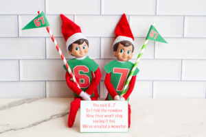 Elf on the Shelf in a green &ldquo;67&rdquo; shirt holding a striped pole with a 67 pennant, standing behind a card that says, &ldquo;You said 6&hellip;7. So I told the reindeer. Now they won&rsquo;t stop saying it too. We&rsquo;ve created a monster.&rdquo;
