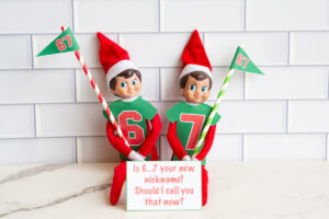 Elf on the Shelf pair wearing printable football-style shirts labeled 6 and 7, carrying 67 flags, displayed with a sign that reads, &ldquo;Is 6&hellip;7 your new nickname? Should I call you that now?&rdquo;