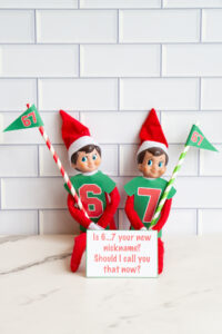 Elf on the Shelf pair wearing printable football-style shirts labeled 6 and 7, carrying 67 flags, displayed with a sign that reads, &ldquo;Is 6&hellip;7 your new nickname? Should I call you that now?&rdquo;