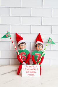 Two Elf on the Shelf dolls dressed in green &ldquo;6&rdquo; and &ldquo;7&rdquo; shirts holding 67 pennant flags, displayed with a printable sign that reads, &ldquo;You said 6&hellip;7. So I tried solving it. Got hungry instead.&rdquo;