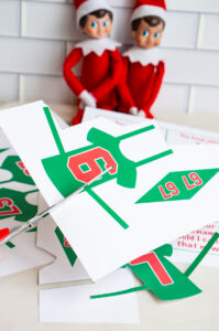 Close-up of hands cutting out a green paper Elf on the Shelf jersey with the number 6 printed on it, with two Elf dolls and additional printable pieces in the background.