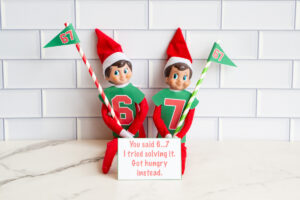 Two Elf on the Shelf dolls dressed in green &ldquo;6&rdquo; and &ldquo;7&rdquo; shirts holding 67 pennant flags, displayed with a printable sign that reads, &ldquo;You said 6&hellip;7. So I tried solving it. Got hungry instead.&rdquo;