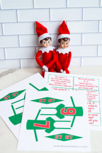 Printable Elf on the Shelf 6&ndash;7 joke set displayed on a table, including cut-out green jerseys with numbers 6 and 7 and matching pennant flags, with two Elf dolls sitting in the background.