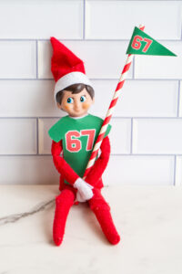 Close-up of a single Elf on the Shelf wearing a green &ldquo;67&rdquo; shirt and holding a matching &ldquo;67&rdquo; flag.