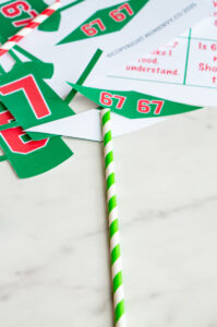 Green and red &ldquo;67&rdquo; printable pennant pieces laid out next to a green-and-white striped paper straw on a marble surface.