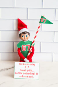 One elf in a green 67 shirt holding a candy-cane-striped 67 flag with a sign reading, &ldquo;You keep saying 6&hellip;7. I don&rsquo;t get it&hellip; but I&rsquo;m pretending I do so I seem cool.&rdquo;