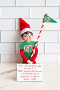 One Elf on the Shelf doll in a green 67 shirt holding a flag, shown with a sign saying &ldquo;Okay&hellip; I keep hearing you whisper 6&hellip;7&hellip; Should I hide??&rdquo;