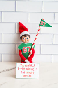 One Elf on the Shelf in a 67 shirt with a matching 67 pennant flag, sitting with a card about getting hungry while solving &ldquo;6&hellip;7.&rdquo;