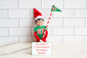 One Elf on the Shelf in a 67 shirt with a matching 67 pennant flag, sitting with a card about getting hungry while solving &ldquo;6&hellip;7.&rdquo;
