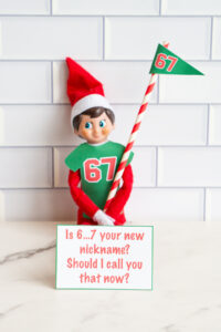 One Elf on the Shelf in a 67 shirt holding a striped flag, posed with a joke card asking if &ldquo;6&hellip;7&rdquo; is the viewer&rsquo;s new nickname.