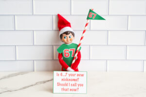 One Elf on the Shelf in a 67 shirt holding a striped flag, posed with a joke card asking if &ldquo;6&hellip;7&rdquo; is the viewer&rsquo;s new nickname.