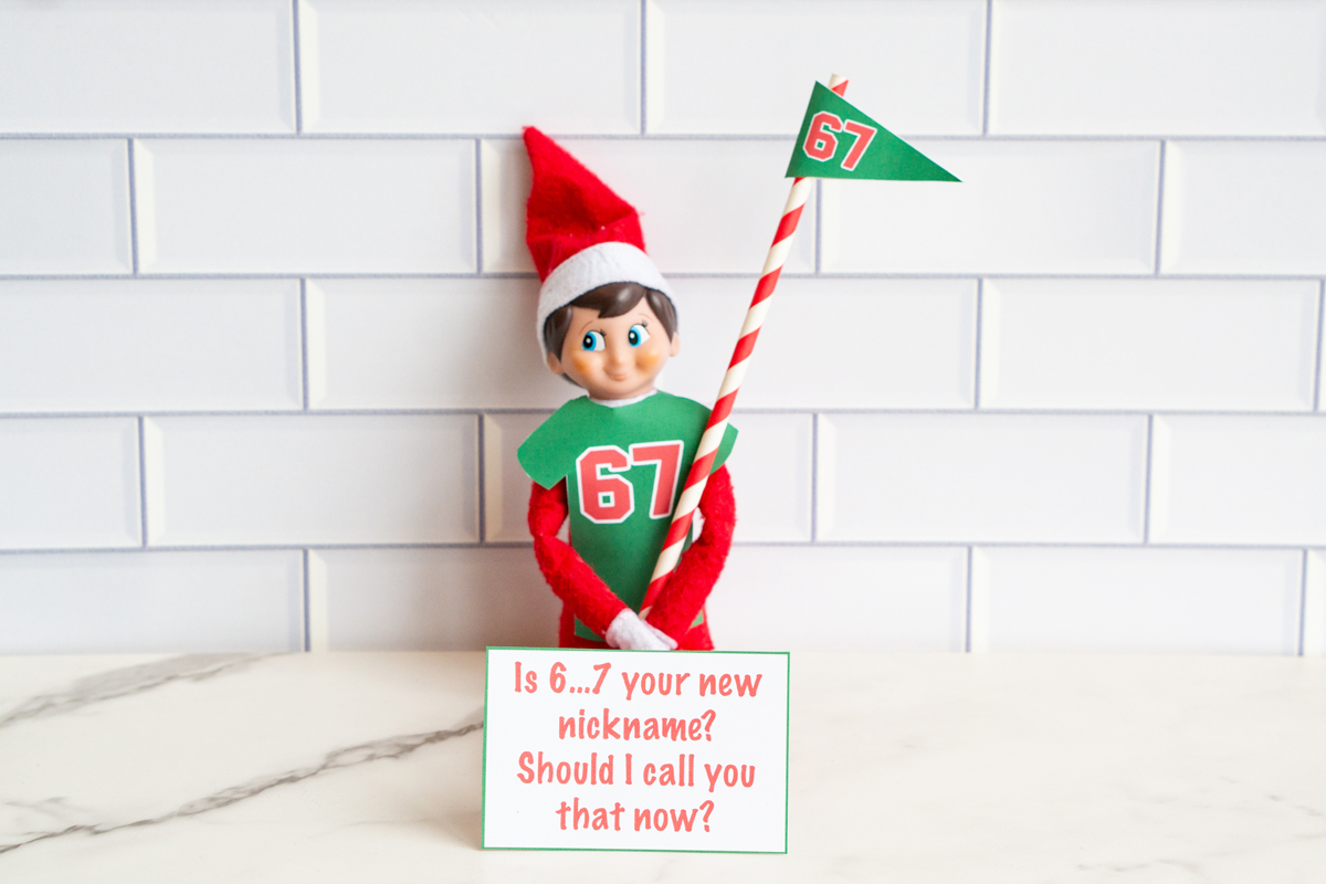 One Elf on the Shelf in a 67 shirt holding a striped flag, posed with a joke card asking if &ldquo;6&hellip;7&rdquo; is the viewer&rsquo;s new nickname.