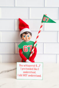 One Elf on the Shelf dressed in a 67 shirt and holding a striped flag, sitting behind a card saying they pretended to understand &ldquo;6&hellip;7&rdquo; but didn&rsquo;t.