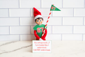 One Elf on the Shelf dressed in a 67 shirt and holding a striped flag, sitting behind a card saying they pretended to understand &ldquo;6&hellip;7&rdquo; but didn&rsquo;t.