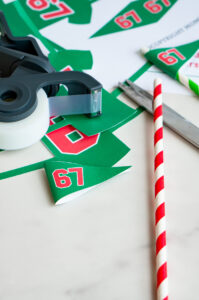 Crafting materials including printed &ldquo;67&rdquo; pennants, scissors, tape dispenser, and a red-and-white striped straw ready for assembling Elf on the Shelf props.