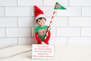 Elf on the Shelf in a green &ldquo;67&rdquo; shirt holding a striped pole with a 67 pennant, standing behind a card that says, &ldquo;You said 6&hellip;7. So I told the reindeer. Now they won&rsquo;t stop saying it too. We&rsquo;ve created a monster.&rdquo;