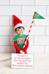 Elf on the Shelf in a green &ldquo;67&rdquo; shirt holding a striped pole with a 67 pennant, standing behind a card that says, &ldquo;You said 6&hellip;7. So I told the reindeer. Now they won&rsquo;t stop saying it too. We&rsquo;ve created a monster.&rdquo;