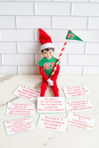 One Elf on the Shelf in a 67 shirt holding a striped 67 flag, sitting alone on a marble surface. The elf has the 12 cut out free printable 6-7 joke cards in front of him laying on the marble.